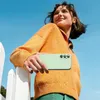 A young woman is holding a green phone case in front of her, leaning against a white fence with a blue sky in the background. Náhled obrázku produktu