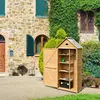 A wooden garden shed with its door open, displaying shelves with gardening tools, set against a stone building with ivy. Produktbild-Vorschau