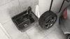A black granite sink and a car tire are placed on a tiled floor next to a car. Náhled obrázku produktu