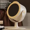 A cream-colored portable heater with a beige accent is shown on a white surface, with a brown fabric background and books in the lower left corner. Produktbild-Vorschau
