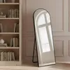A tall, arched mirror stands on a wooden floor next to a wooden bookshelf filled with books. The mirror reflects a window with decorative panes. Produktbild-Vorschau