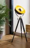A black tripod floor lamp with a gold interior is placed next to a large potted plant on a wooden floor. Podgląd zdjęcia produktu