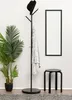 A black stool is placed next to a coat rack with a hat and a blanket on it, in front of a wall with a framed picture and a plant. Náhled obrázku produktu