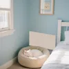 A white cat sleeps in a beige cat bed next to a bed in a room with light blue walls. Produktbild-Vorschau