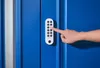 A hand is shown pressing buttons on a white keypad attached to a blue door. Anteprima dell’immagine prodotto