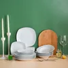 A collection of white dinnerware, including plates and bowls, is arranged on a beige surface against a green background. Two white candlesticks with tall, twisted candles stand to the left, and a decorative wooden arch is placed behind the dinnerware. A wi Náhled obrázku produktu