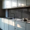 A modern kitchen with light gray cabinets and a dark herringbone backsplash. Sunlight streams in, casting shadows. Produktbild-Vorschau