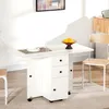 A white drop-leaf table with drawers and storage cabinets, set up in a room with a chair and a framed picture on the wall. Produktbild-Vorschau