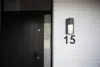 A modern black front door with a black handle and a small peephole, next to a white brick wall with a black outdoor light fixture and the house number "15" in black. Anteprima dell’immagine prodotto