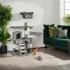 A cat tree is shown in a living room setting with a sofa and plants. Náhled obrázku produktu