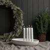 A white ceramic candle holder with four white candles is placed on a gray linen cloth. A Christmas wreath and a small evergreen plant are visible in the background. Produktbild-Vorschau