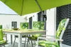 An outdoor dining set with chairs and a table, covered by a large green umbrella. The chairs and cushions have a tropical palm leaf pattern. Produktbild-Vorschau