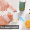 A hand is pressing a button on a white mini vacuum machine, which is attached to a vacuum seal bag containing chicken. A green mini vacuum machine and a lemon slice are also visible in the background. Produktbild-Vorschau
