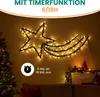 A decorative star-shaped light fixture with trailing lights, resembling a shooting star, is displayed on a wall. A Christmas tree is visible on the left, and a mantelpiece with Christmas decorations is on the right. The scene is set indoors, likely during  Produktbild-Vorschau