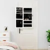 A white jewelry cabinet with mirrors is mounted on a white door, open to reveal organized jewelry. A dresser and a bed are partially visible to the left. Voir les images du produit