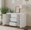 A white sideboard with three drawers and two doors, placed in a room with a plant and a lamp. Náhled obrázku produktu