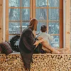 A woman and a child are sitting on a wooden bench by a window, looking out at the winter scenery. The window has Christmas tree decals on it. Produktbild-Vorschau