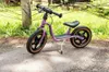 A purple balance bike is parked on a gravel path with green grass and a wooden post in the background. Voir les images du produit