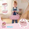 A young girl is holding a doll carrier with dolls inside, in a room with a teepee and shelves. Produktbild-Vorschau