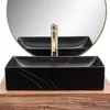 A black rectangular vessel sink with gold veining sits on a wooden countertop, with a gold faucet and a round mirror in the background. Produktbild-Vorschau