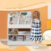 A young girl stands in front of a white bookshelf filled with books and toys, in a room with an orange wall. Produktbild-Vorschau