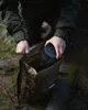 A person is putting a black cylindrical object into a brown backpack in a forest setting. Produktbild-Vorschau