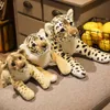 Three plush toy wild cats (lion cub, tiger cub, and leopard) are lying on a textured surface, possibly a sofa or rug. In the background, a basket of yellow fruit is visible. Náhled obrázku produktu