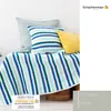 A knitted blanket and pillows are arranged on a wooden chest against a white wall. Produktbild-Vorschau