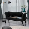 A black storage bench with its lid open, revealing stored items, in a living room setting with a floor lamp and a side table with decorations. Podgląd zdjęcia produktu
