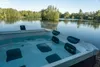 A hot tub filled with water, situated next to a calm lake surrounded by trees. Náhled obrázku produktu
