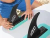 A person is attaching fins to a paddleboard on a beach. Produktbild-Vorschau