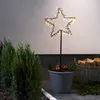 A star-shaped garden stake with LED lights is placed in a pot with red apples and greenery, next to a basket of firewood and evergreen trees. Podgląd zdjęcia produktu