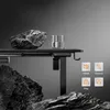 A black adjustable desk is shown with a large rock and a glass of water on it, with other rocks in the foreground and background. Icons indicating features are displayed on the right. Anteprima dell’immagine prodotto