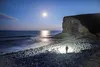 A person stands on a rocky beach at night, illuminated by a flashlight, with the moon and ocean in the background. Produktbild-Vorschau