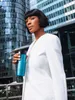 A woman in a white blazer holds a teal travel mug in front of a modern glass building. Produktbild-Vorschau