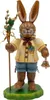 Ein fröhlicher Hase in traditioneller Kleidung hält einen Stab mit Blumen in der einen Hand und eine Glocke in der anderen während er auf einem grünen Sockel steht und freundlich lächelt Produktbild-Vorschau