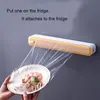 A person is using a plastic wrap dispenser attached to a refrigerator to cover a plate of food. Produktbild-Vorschau