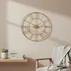 A gold wall clock with Roman numerals hangs on a light-colored wall above a small wooden shelf with books and a potted plant, next to a chair with a blanket. Voir les images du produit