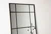 A close-up of a black framed mirror with a grid pattern, leaning against a white wall. A reflection of dried flowers is visible in the mirror. Podgląd zdjęcia produktu