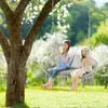 Two young girls are sitting on a double hanging chair in a garden setting. Voir les images du produit