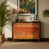 A vintage-style sideboard with oriental Chinese Asian design elements, featuring intricate circular patterns on the doors and drawers, placed in a room with a plant and artwork. Produktbild-Vorschau