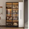 A modern display cabinet with glass doors and internal LED lighting, showcasing various items like bottles, dishes, and decorative objects. It is placed in a room with a neutral wall and floor. Podgląd zdjęcia produktu