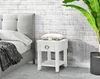 A bedside table next to a bed, with a plant in the background. Náhled obrázku produktu