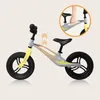 A balance bike with a gray and yellow frame, black wheels and seat, shown on a light beige background. An inset circle shows a hand holding the handlebars. Produktbild-Vorschau