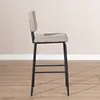 A side view of a taupe bar stool with a black metal frame, set against a light beige wall and a wooden floor. Produktbild-Vorschau
