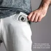 A person's hand is placing a compact digital camera into the pocket of white jeans. Produktbild-Vorschau