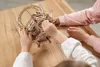 Two people, likely a child and an adult, are assembling a wooden mechanical puzzle on a wooden table. Podgląd zdjęcia produktu