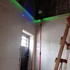 A laser level mounted on a telescopic pole is set up in a room with tiled walls and a dark ceiling, possibly during construction or renovation. Náhled obrázku produktu