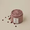 A jar of pinkish-red pore mask with red beans scattered around it on a light beige background. Produktbild-Vorschau