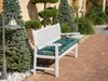 A white garden bench with a patterned cushion sits on a brick patio surrounded by lush greenery and a lamppost. Náhled obrázku produktu